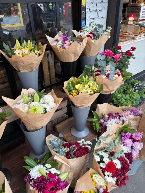 A display of various fresh flower bouquets arranged in tall, tapered, dark grey vases and wrapped in brown craft paper. The flowers include white lilies, yellow daisies, purple statice, pink carnation