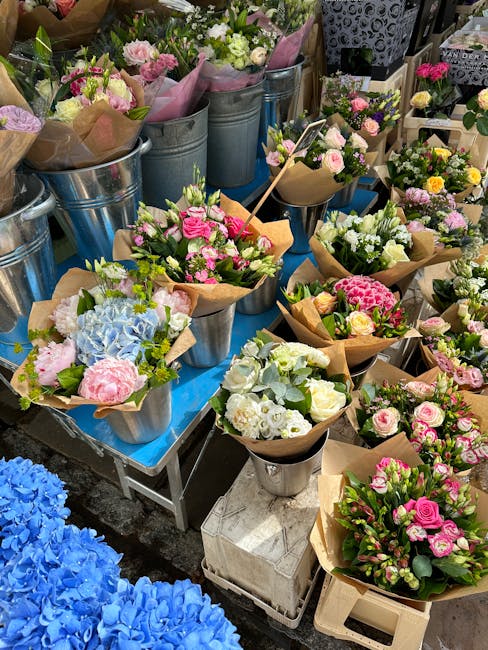 A display of various fresh floral bouquets arranged in metal buckets and wrapped in brown craft paper, featuring pink, white, and pastel-colored roses, hydrangeas, and lisianthus. The bouquets are set