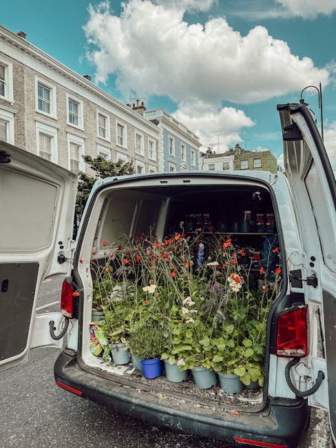 The open back of a white delivery van revealing a variety of potted flowers, including red poppies, white daisies, and green leafy plants, arranged in light blue pots and placed on a shelf inside the