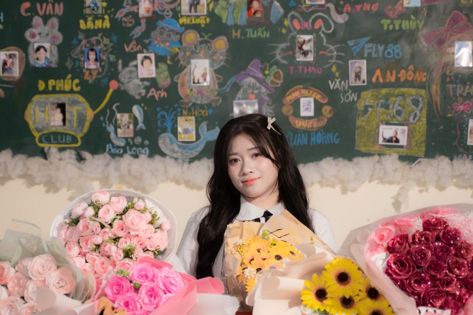A young woman with long black hair, styled with a small silver hair clip, stands behind a table arranged with various floral bouquets. She has a gentle smile and is wearing a white blouse with a black