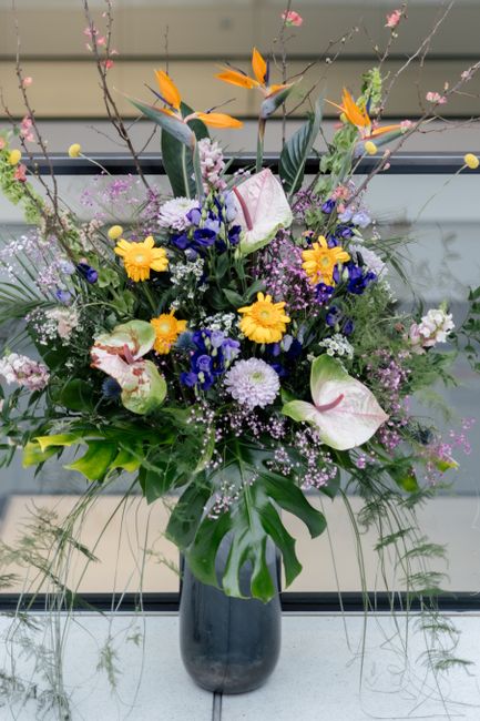 A vibrant floral arrangement displayed in a tall, dark glass vase, featuring a mix of fresh flowers including large white calla lilies with pink streaks, bright yellow daisies, deep blue lobelias, and